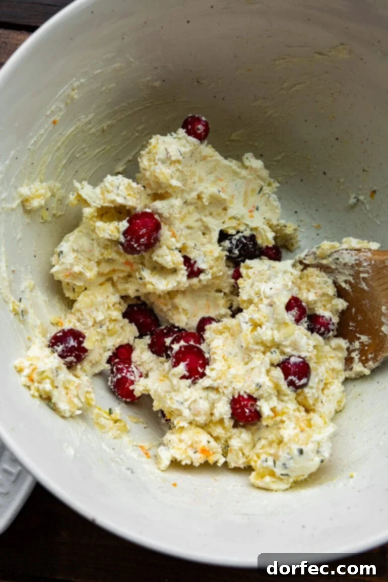 a photo of cream cheese, whole cranberries and grated cheese being stirred together in a white bowl with a wooden spoon