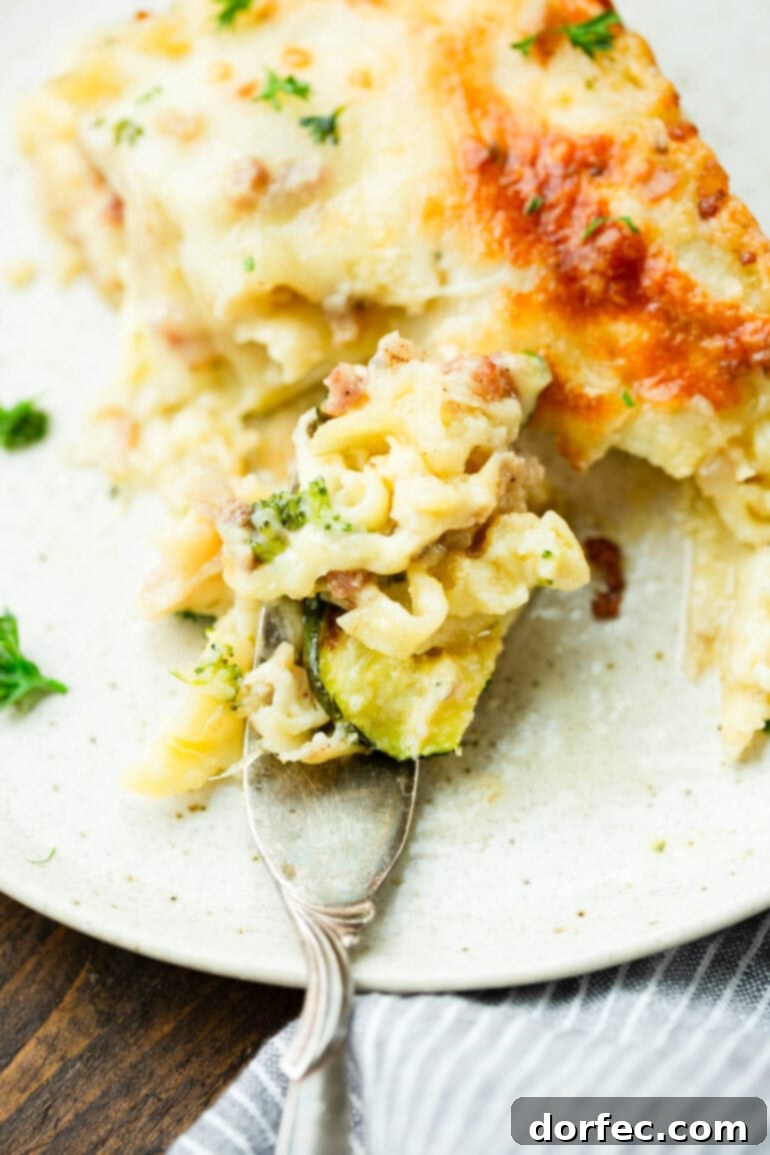A photo of a slice of pancetta vegetable lasagna with white sauce being cut up and eaten with a fork, illustrating its inviting texture and deliciousness.
