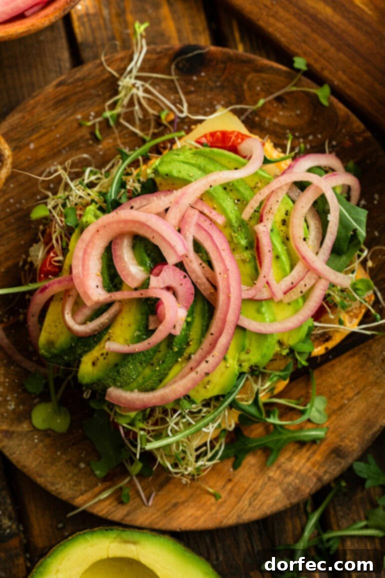 An open avocado sandwich displaying all its delicious layers: creamy avocado slices, vibrant pickled red onions, fresh bean sprouts and microgreens, rich Manchego cheese, and charred cherry tomatoes.