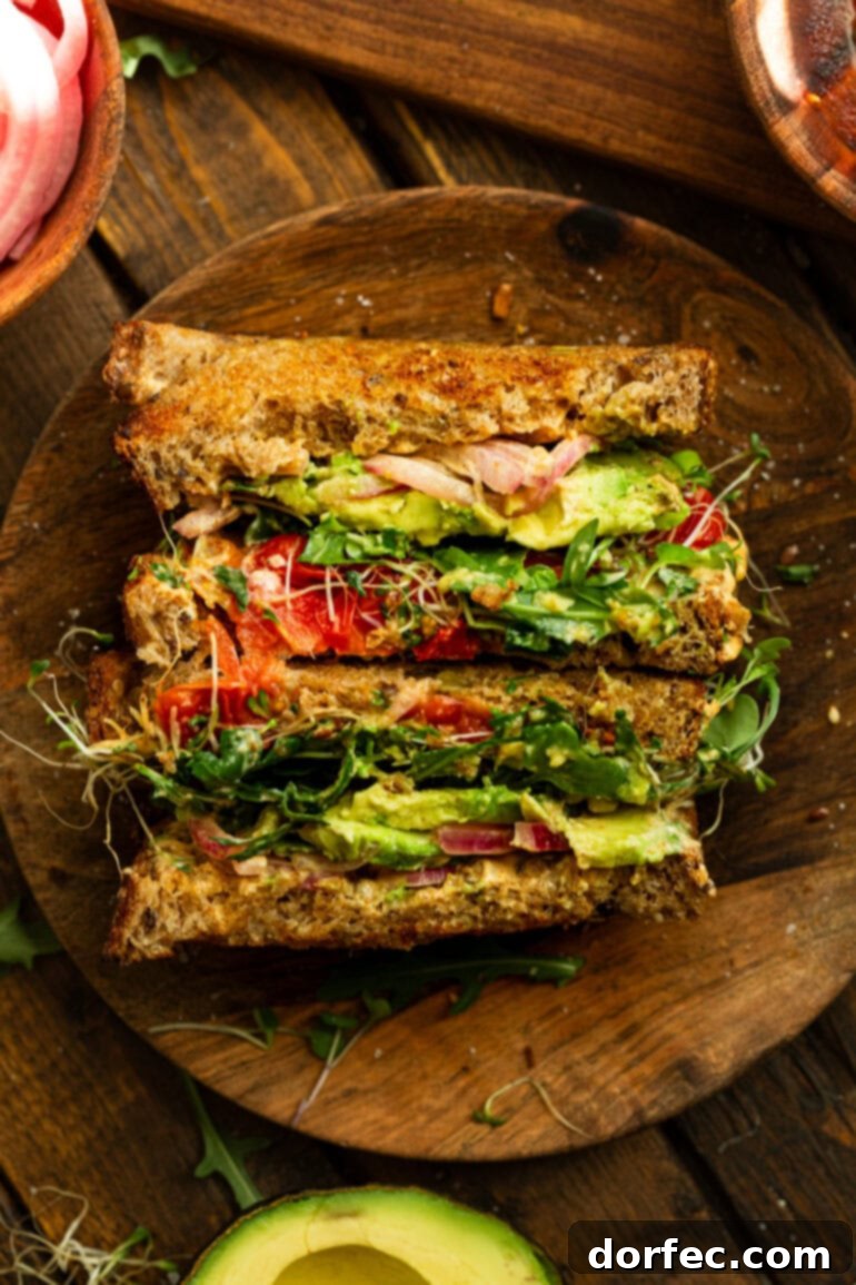 A beautifully presented avocado sandwich, sliced in half and arranged to show off its enticing layers of fresh ingredients, including avocado, pickled red onions, sprouts, and cheese.