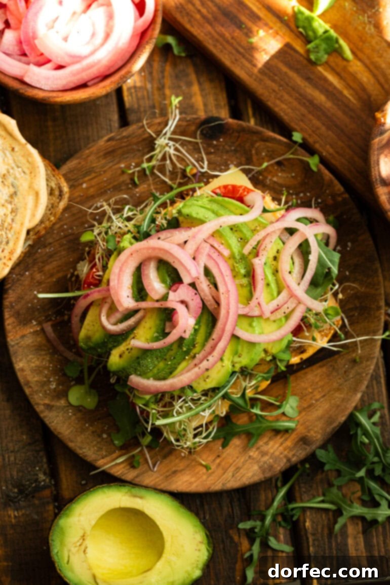 A close-up view of an open avocado sandwich, revealing perfect slices of avocado, vibrant pickled red onions, and a generous layer of microgreens and bean sprouts, all nestled on toasted bread.