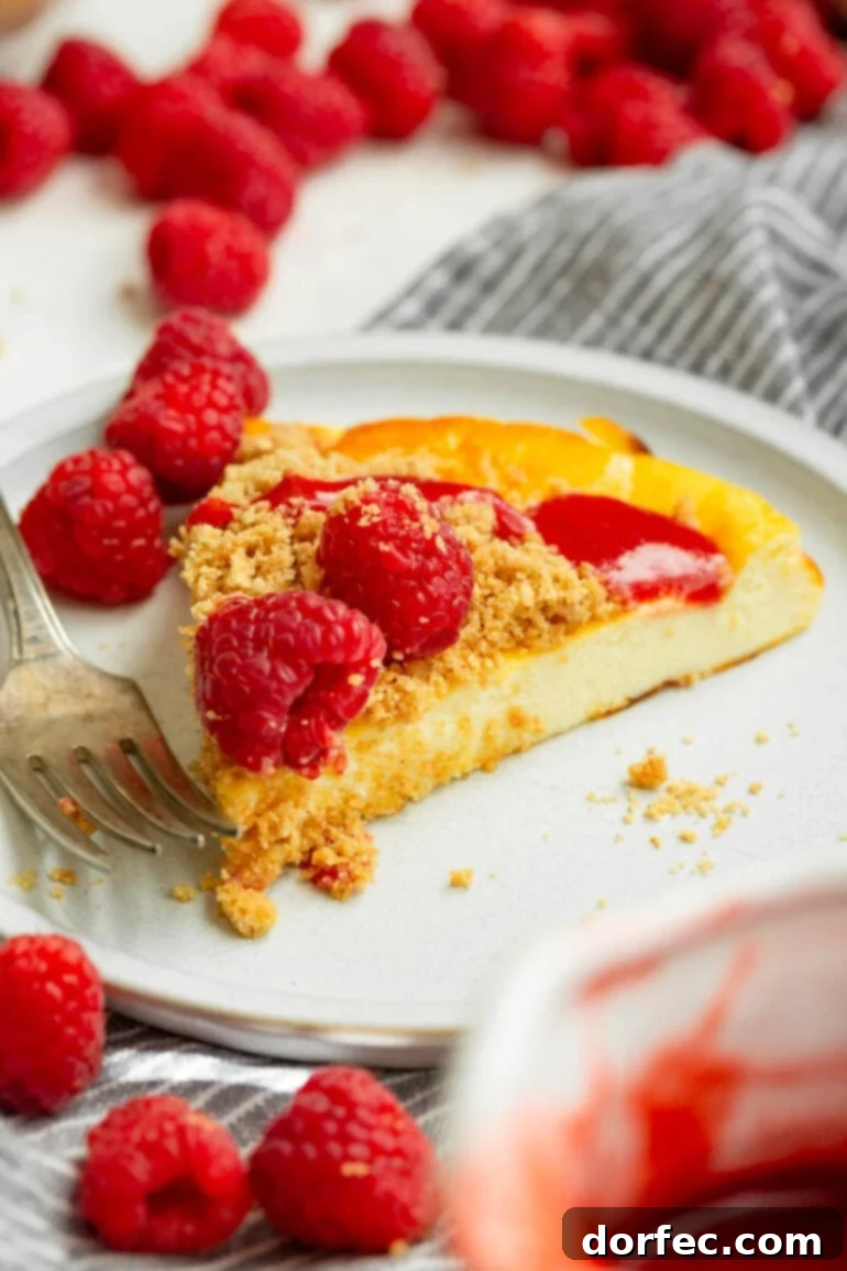 Creamy Baked Yogurt Cheesecake 6 A photo of a slice of baked yogurt on a small white plate, topped with whole raspberries, graham cracker crumb topping, and a raspberry drizzle.