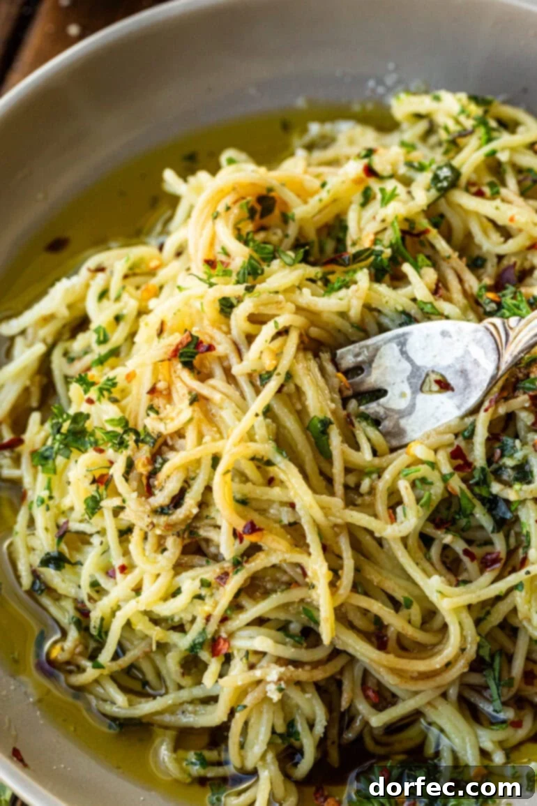 A fork twirling cooked spaghetti pasta, glistening with olive oil and speckled with fresh herbs and garlic pieces.