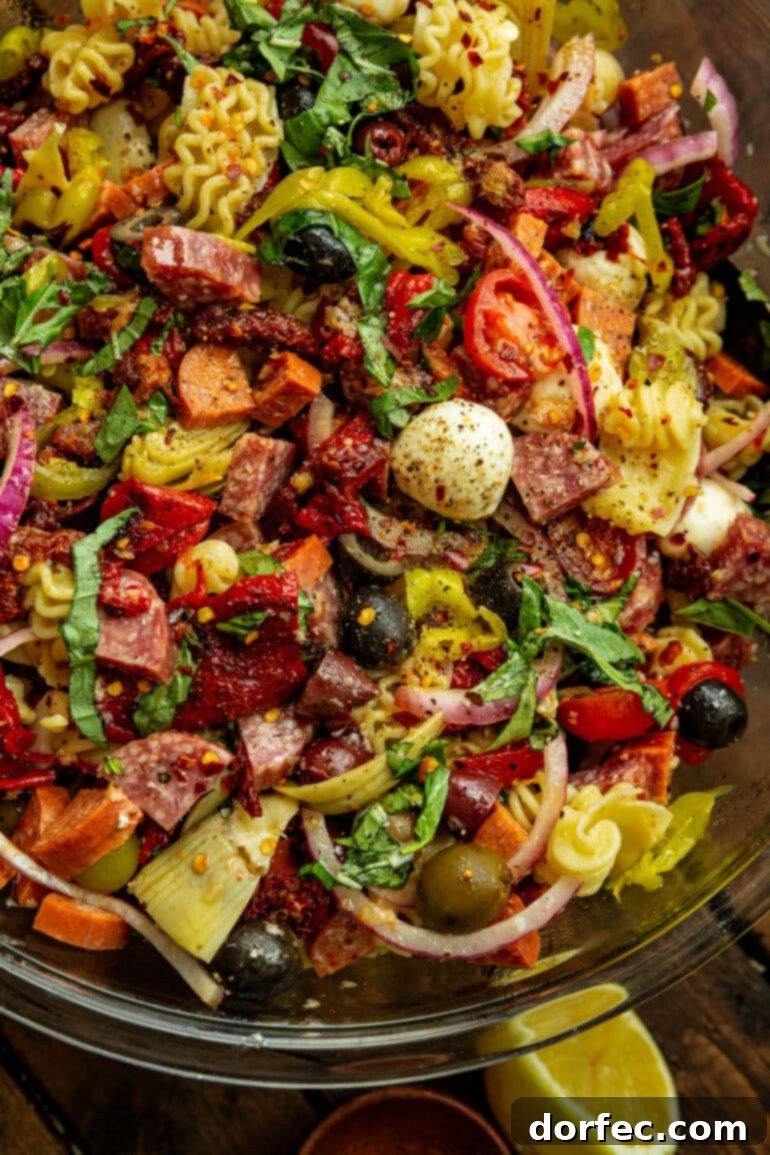 A vibrant antipasto pasta salad in a large glass bowl, showcasing cooked rotini pasta, mozzarella pearls, pepperoncinis, salami, pepperoni, and olives.