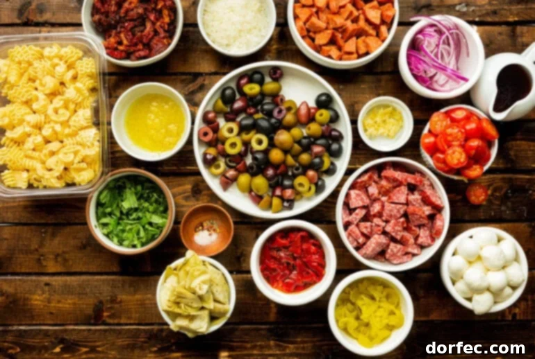 An overhead view of all the prepared ingredients for antipasto pasta salad, neatly arranged in separate small bowls, including olives, salami, pepperoni, fresh basil, artichoke hearts, and roasted red peppers.
