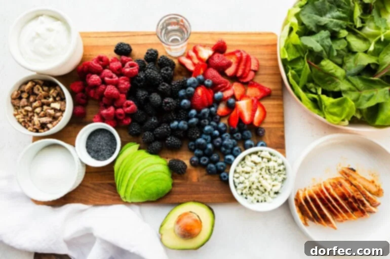 A beautifully arranged flat lay photo of all the fresh ingredients for Berry Avocado Chicken Salad, including colorful berries, creamy avocado, tangy blue cheese, crisp lettuce, and the dressing components.