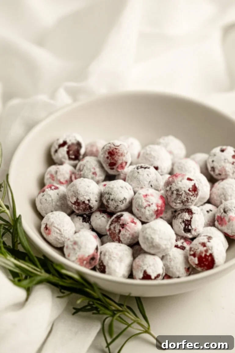 Glittering Sweet Tart Cranberries 2 A small bowl overflowing with sparkling candied cranberries, garnished with a sprig of fresh rosemary, resting on a white surface.