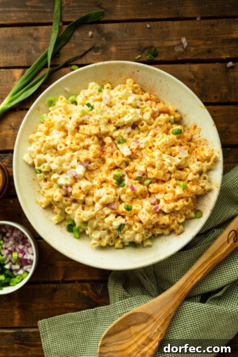 A beautifully presented large white bowl of creamy deviled egg pasta salad, garnished with fresh green onions and smoked paprika, surrounded by a wooden spoon and other garnishes.