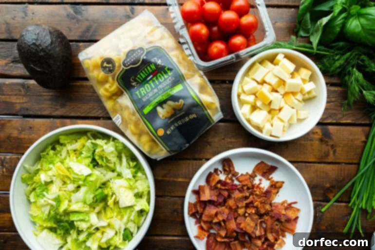 a photo of all the ingredients for blt pasta salad in separate bowls including bacon crumbles, cubed cheese, chopped lettuce, a bag of pasta and cherry tomatoes