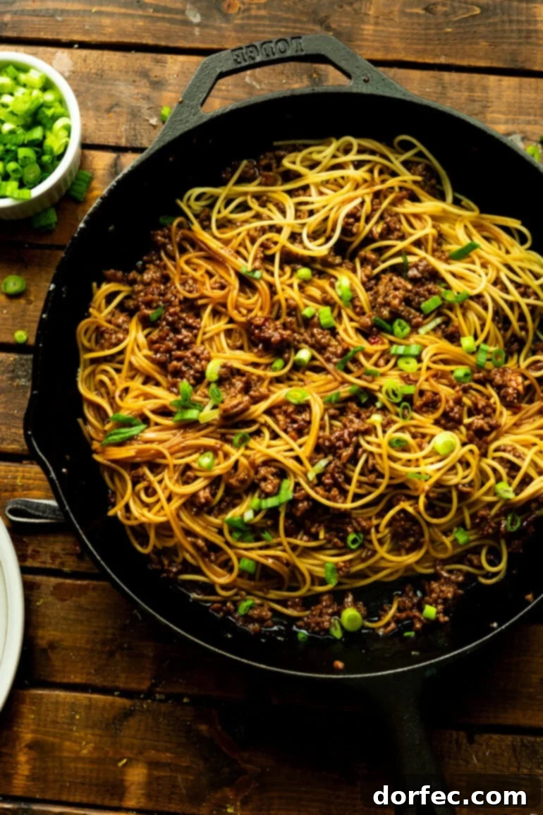 An overhead shot of a cast iron skillet brimming with cooked spaghetti noodles perfectly coated in a savory ground beef sauce, topped with vibrant chopped scallions, ready to be served family-style.