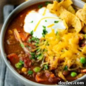 A vibrant bowl of hearty taco stew, generously topped with a dollop of cool sour cream, a mound of shredded cheddar cheese and crispy Fritos chips.