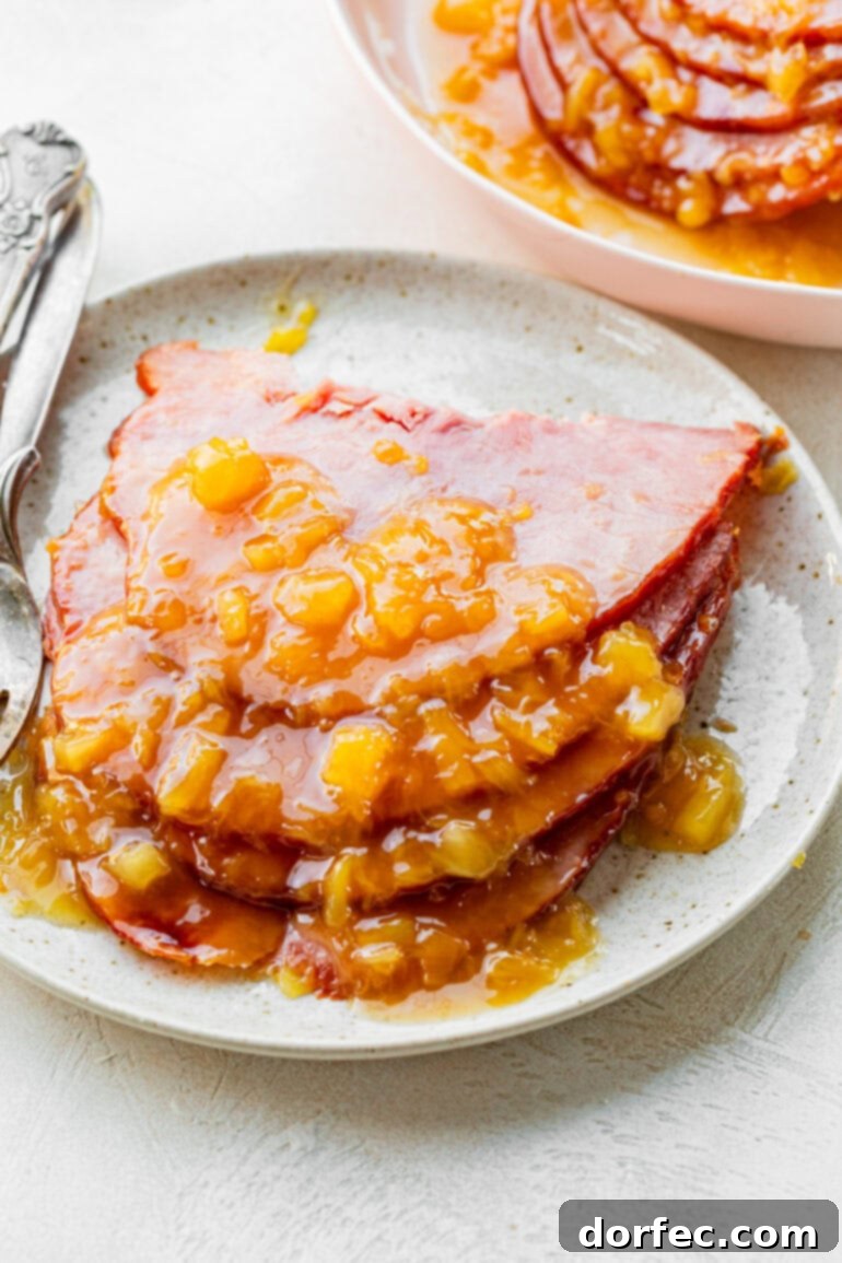 Perfectly Tender Slow Cooker Ham 8 A close-up photo of several succulent slices of slow cooker ham, neatly stacked on a pristine white plate and lavishly topped with a glistening pineapple brown sugar glaze.