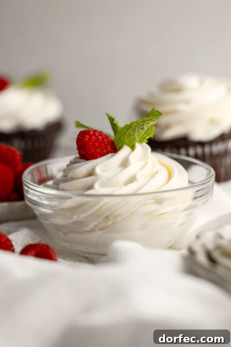 Fluffy Tangy Cream Cheese Frosting 10 A charming glass bowl brimming with beautifully piped whipped cream cream cheese frosting, elegantly garnished with a fresh raspberry and a few vibrant mint leaves, suggesting gourmet delight.
