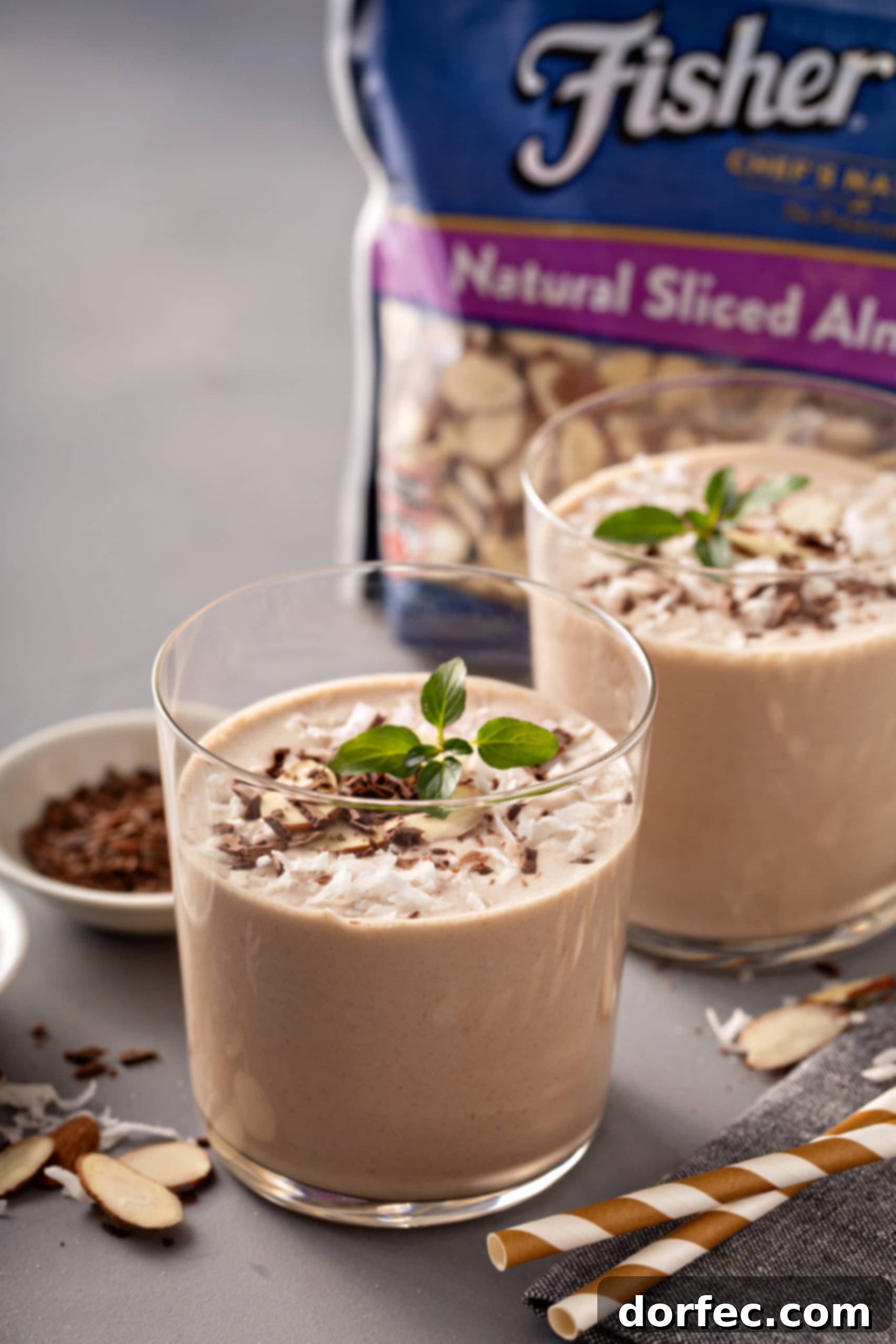 Almond Joy Protein Boost 3 Two glasses of Almond Joy Protein shakes, elegantly topped with shaved chocolate, fresh coconut flakes, and slivered almonds, accompanied by a package of Fisher's slivered almonds and mint leaves for garnish.