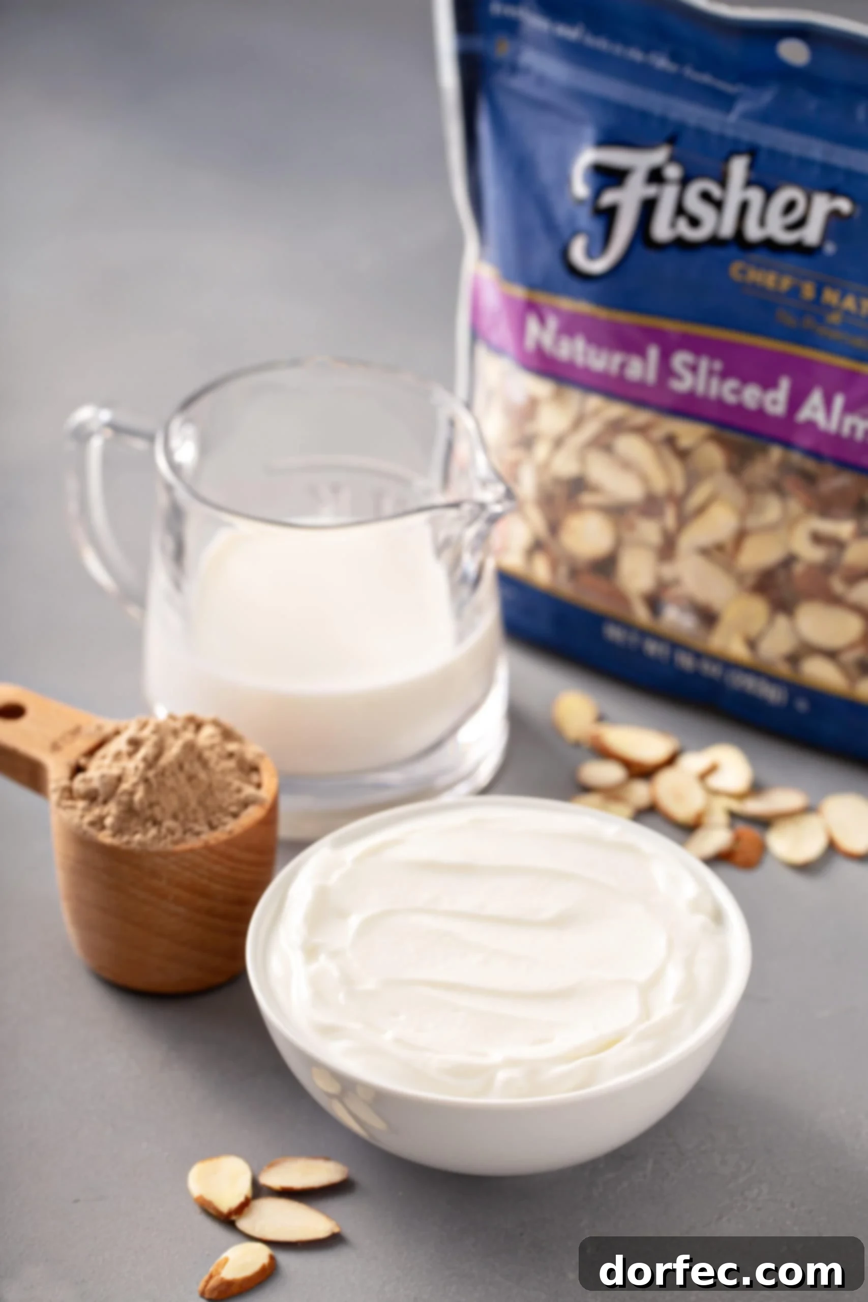 Almond Joy Protein Boost 5 Collection of fresh ingredients arranged for an Almond Joy Protein Shake: a tub of creamy yogurt, a carton of milk, a scoop of chocolate protein powder, and a generous portion of whole almonds.