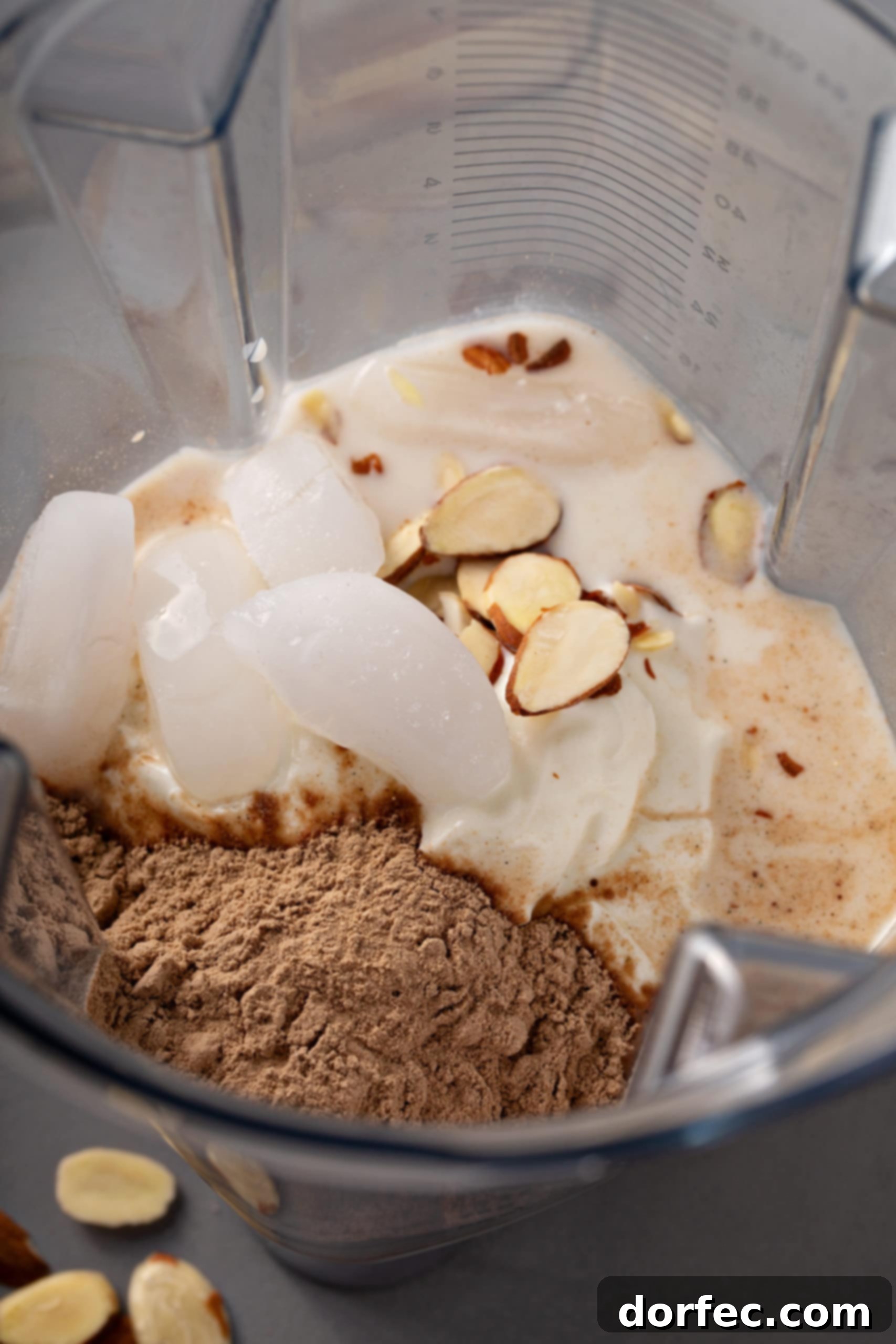 Almond Joy Protein Boost 9 Blender jar filled with ingredients for a delicious chocolate coconut protein shake, including ice cubes, whole almonds, rich chocolate protein powder, creamy yogurt, and a splash of milk.