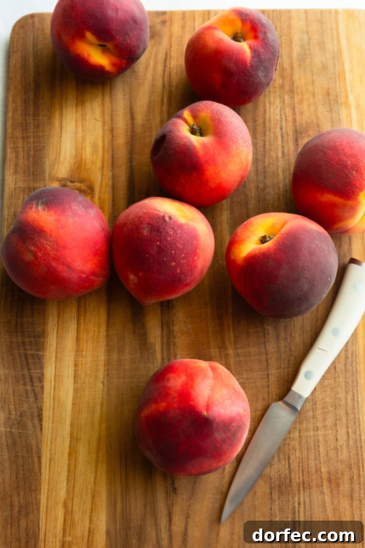 Whole ripe peaches resting on a wooden cutting board next to a paring knife.