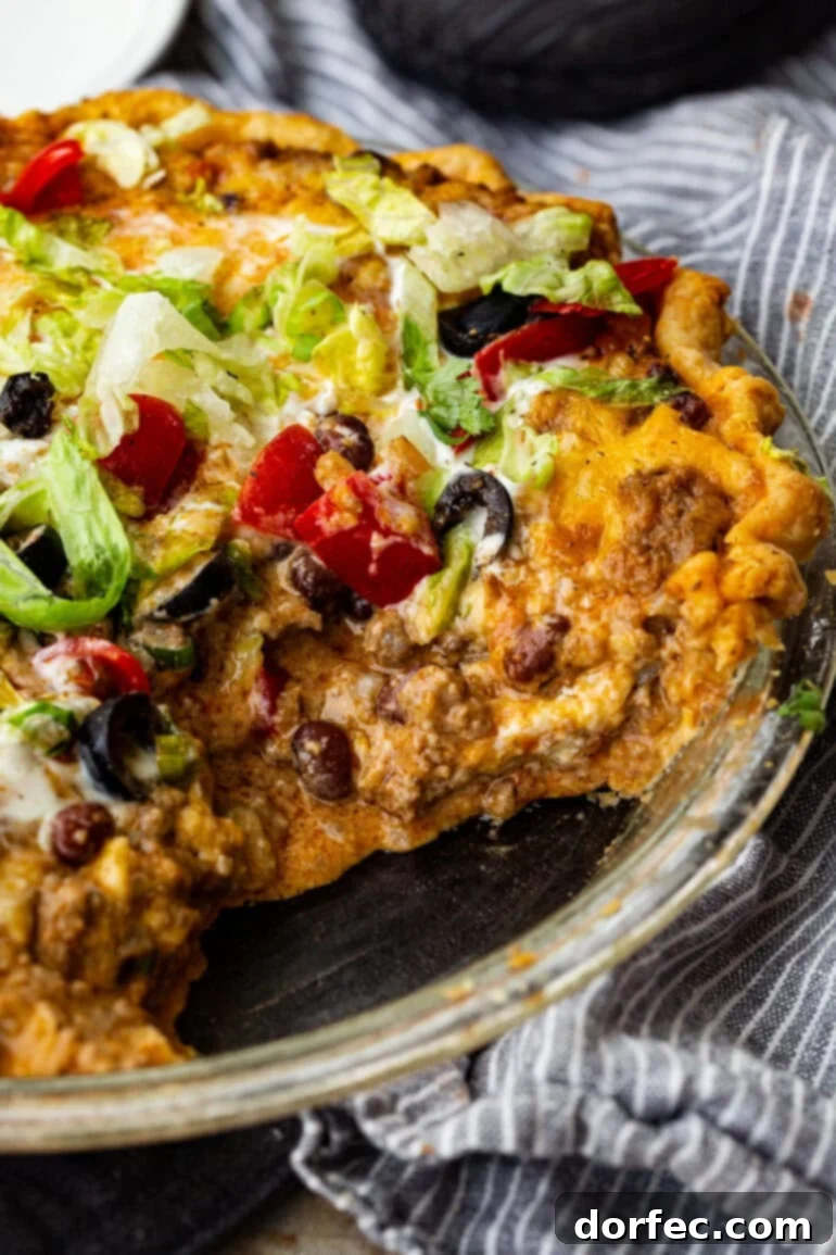 A vibrant Taco Pie in a pie tin, generously topped with melted cheese, fresh shredded lettuce, diced tomatoes, a dollop of sour cream, and sliced black olives. Ready to serve, highlighting its delicious layers.