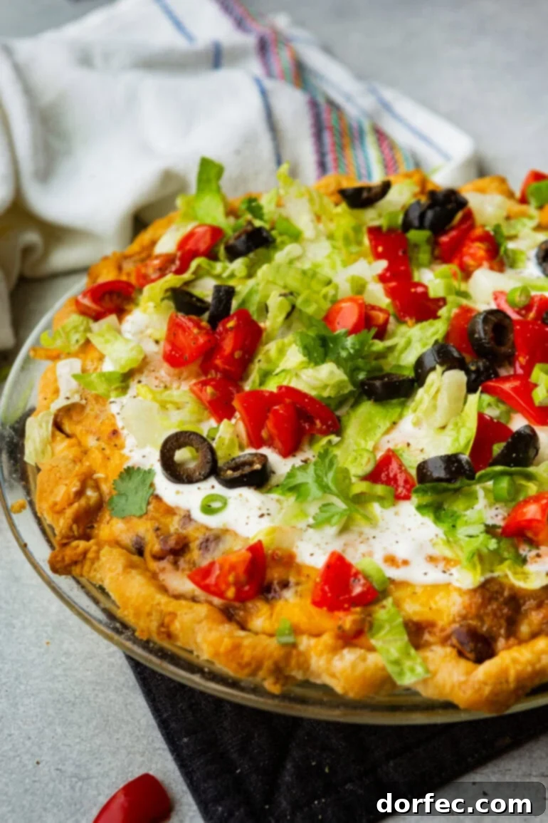 A perfectly baked whole Taco Pie sitting in a glass pie plate, generously adorned with a vibrant array of fresh toppings: creamy sour cream, crisp shredded lettuce, bright diced tomatoes, and savory sliced black olives. A true crowd-pleaser and easy weeknight dinner.