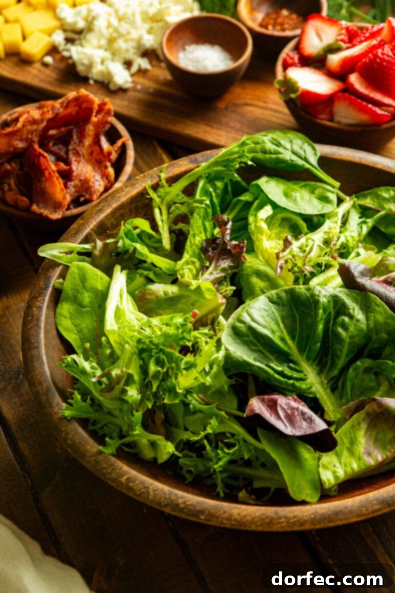 A top-down view of a large wooden serving bowl filled with vibrant mixed greens, with a smaller bowl of perfectly cooked, crumbled bacon in the background, ready to be added to the salad.
