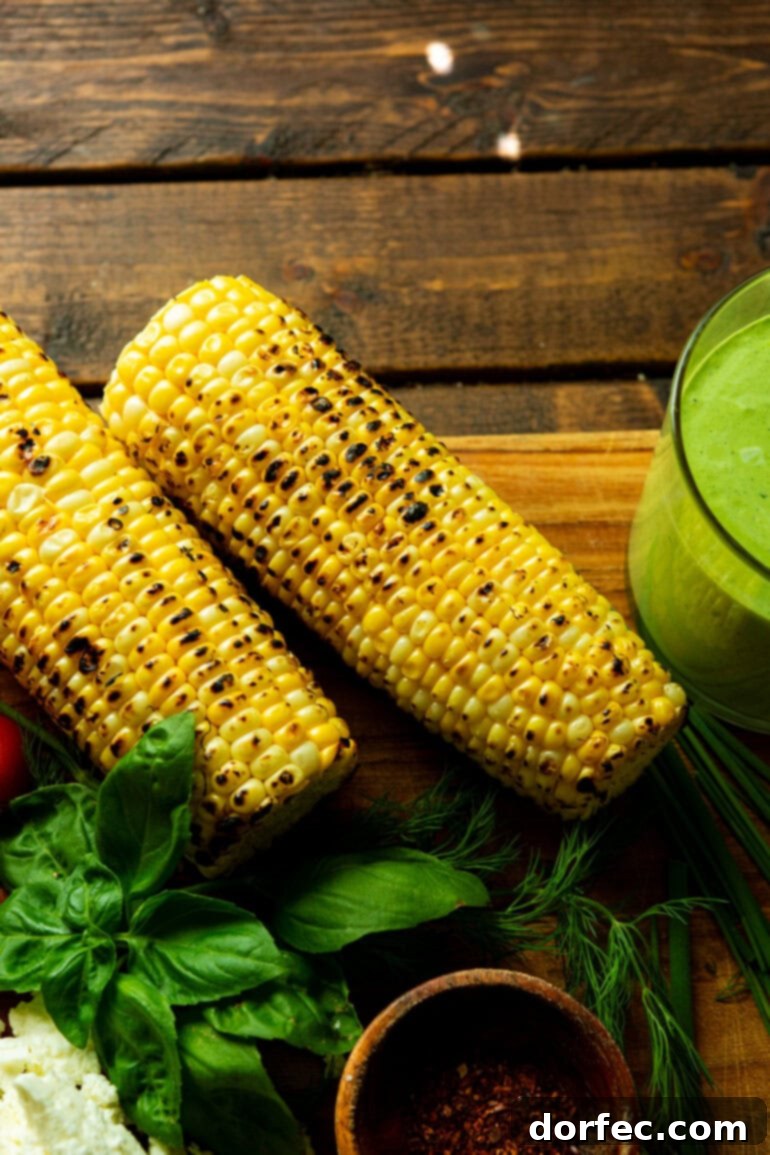 A close-up of two perfectly charred corn on the cobs resting on a wooden board, with a glass filled with creamy basil dressing next to them, showcasing key salad components.