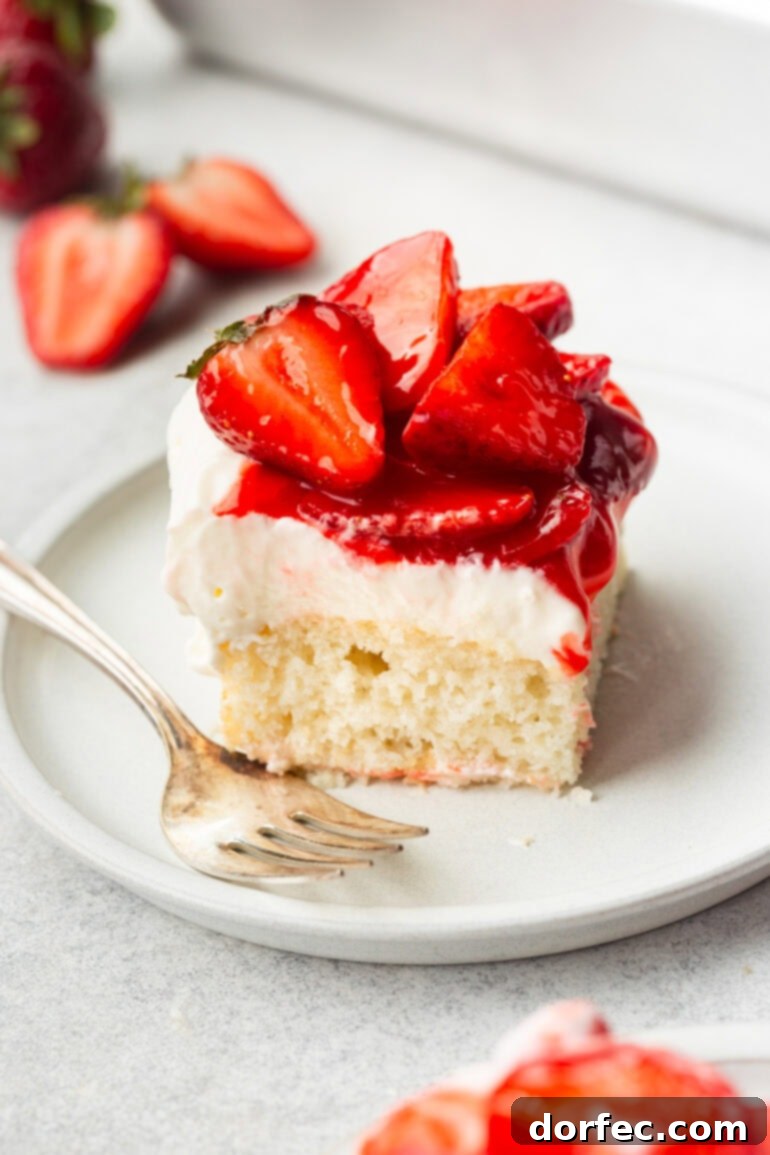 A pristine slice of strawberry shortcake cake on a small white dessert plate, featuring perfectly layered moist white cake, creamy frosting, and saucy halved strawberries, ready to be enjoyed.