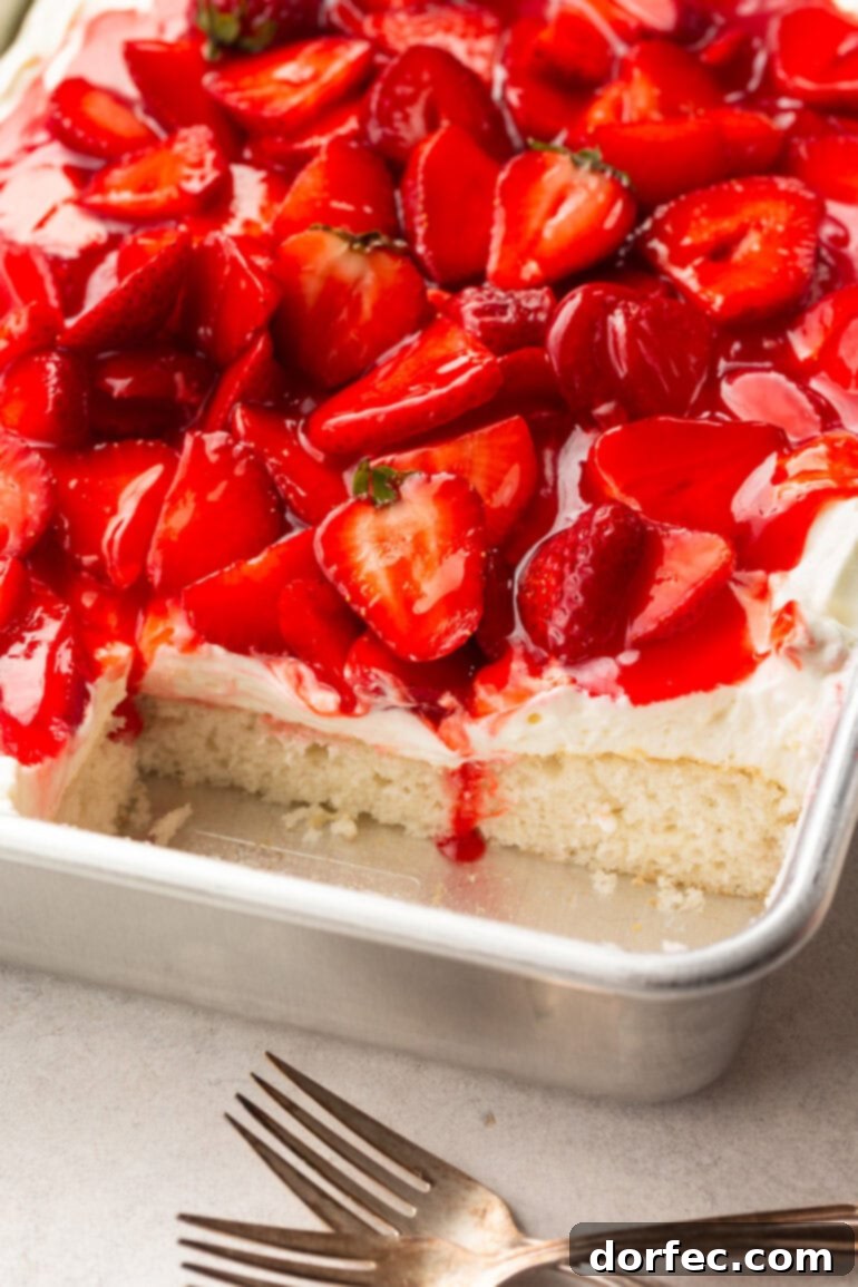 A whole strawberry shortcake cake in a 9x13 metal cake pan, with a piece expertly removed to showcase the cross-section of moist cake, fluffy cream, and luscious strawberries.