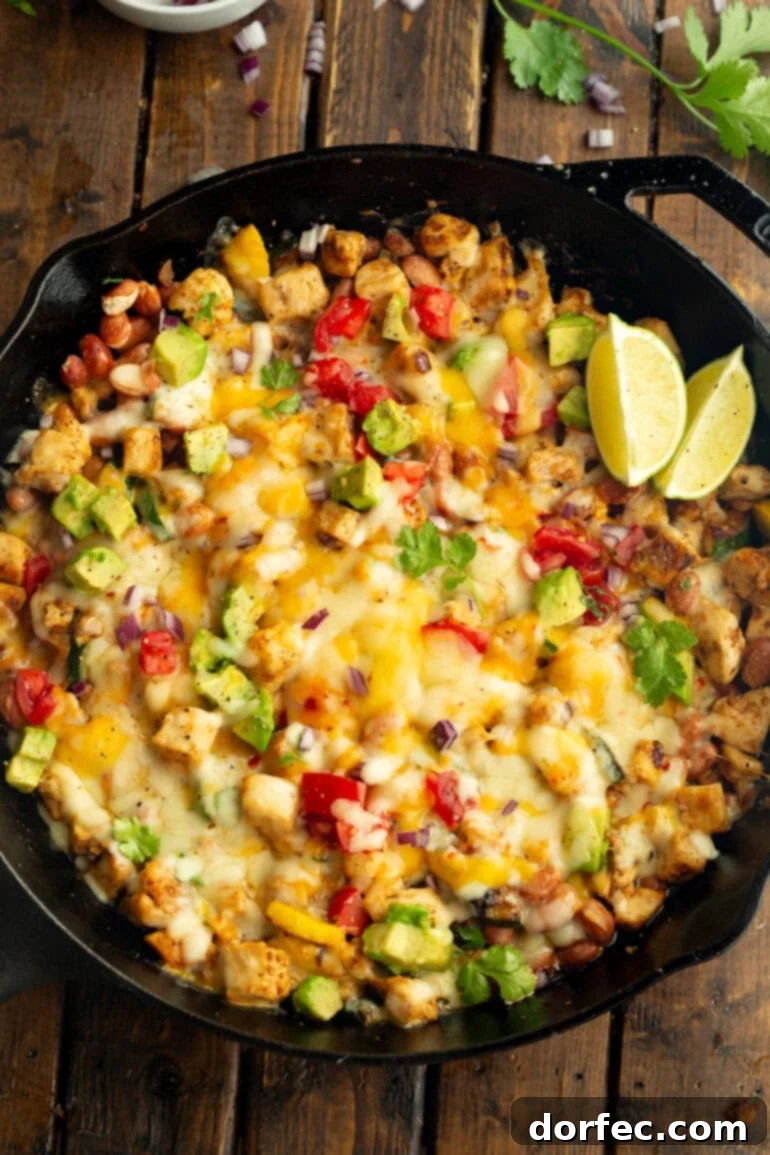 A cast iron skillet overflowing with golden-brown chicken chunks, vibrant melted cheese, fresh avocado slices, tender zucchini, and zesty lime wedges, ready to serve.