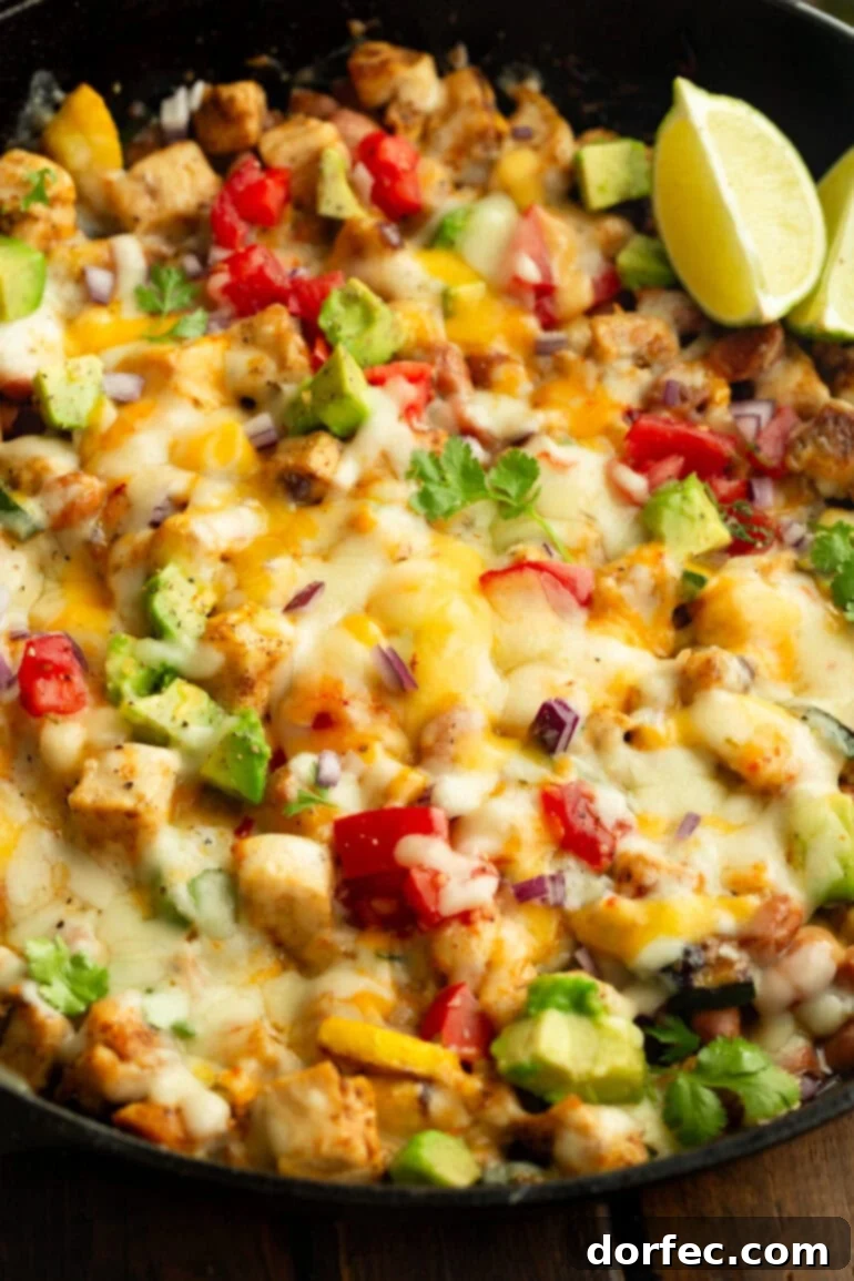 A close-up view of the melted cheese generously covering tender chicken pieces, diced tomatoes, fresh zucchini, and creamy avocado in a cast iron skillet.
