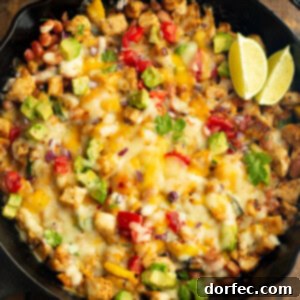 a cast iron skillet full of golden chicken chunks, melted cheese, avocado, zucchini and a couple of lime wedges