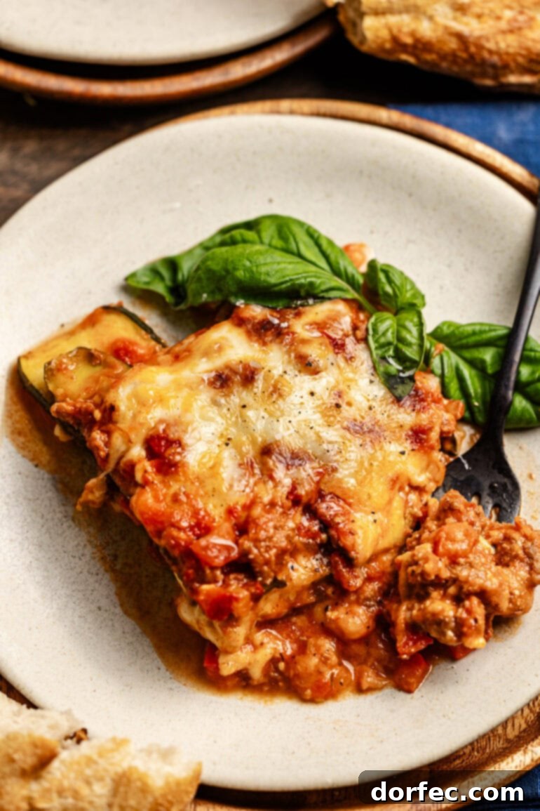 a plate with a hearty slice of layered zucchini noodles, cheese, and meat sauce with a basil garnish. A fork is digging into the slice of lasagna.