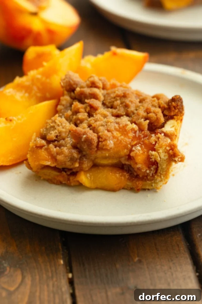 A slice of peach crumble bar on a white plate, topped with golden-brown streusel and served with fresh peach slices. This delectable dessert features a flaky crust, juicy peaches, and a rich, buttery brown sugar crumble, making it an irresistible summer treat.
