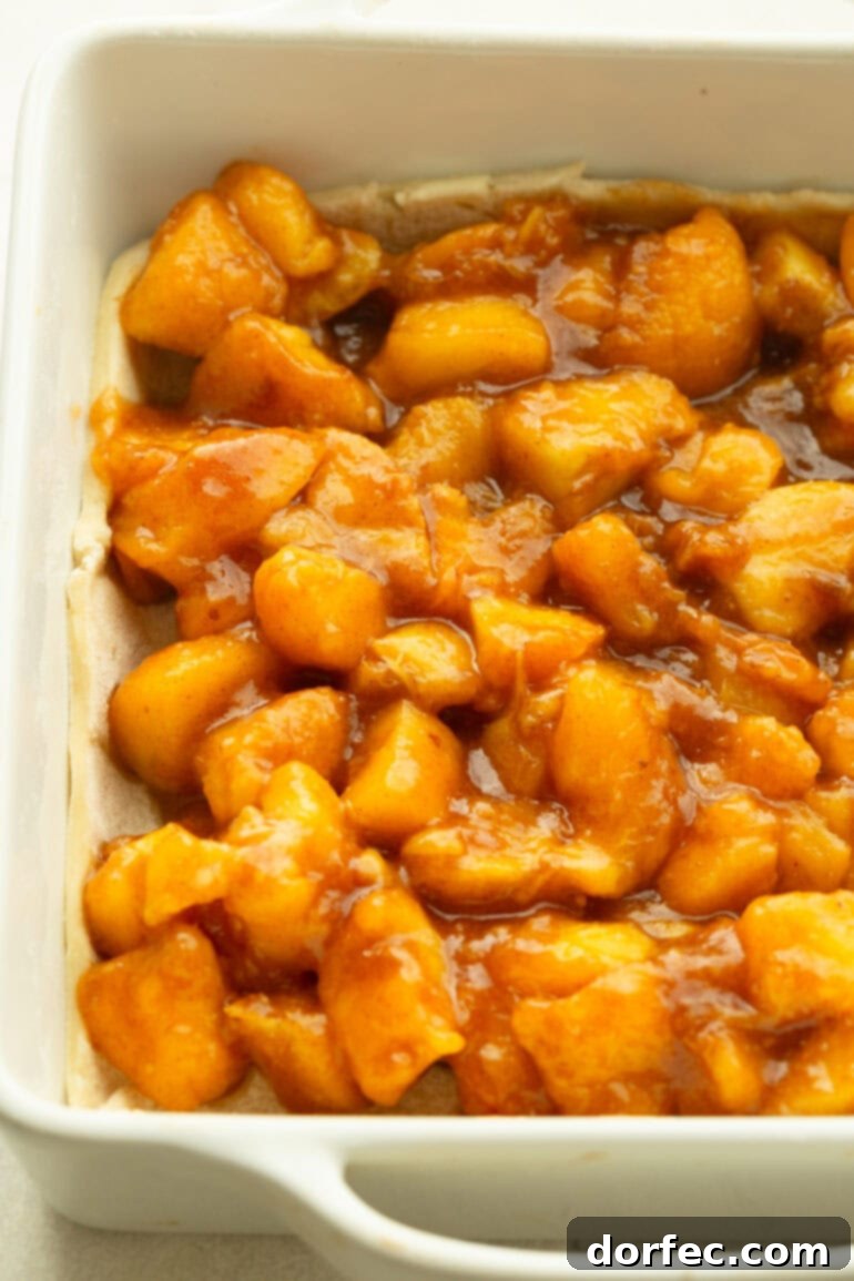 Close-up of baked peach filling in a white baking dish, showing soft, juicy peach chunks coated in a glossy cinnamon-sugar sauce, ready to be topped with streusel.