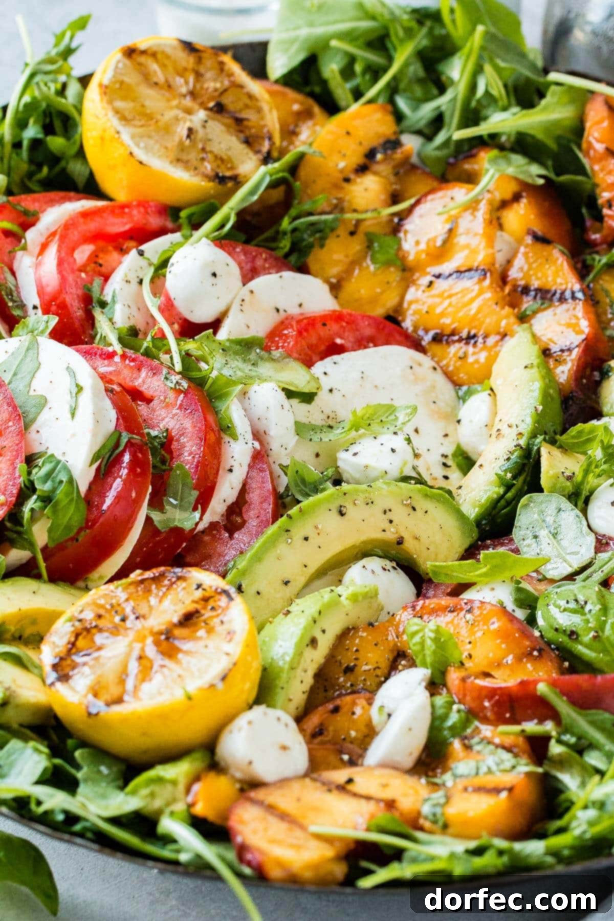 Sweet and Savory Grilled Peach Caprese 5 Close up of Griled Peach Caprese Salad filled wiht slices of avocado, tomato, mozzarella, green leaves, grilled peaches, and two halves of lemons coated with balsamic glaze.