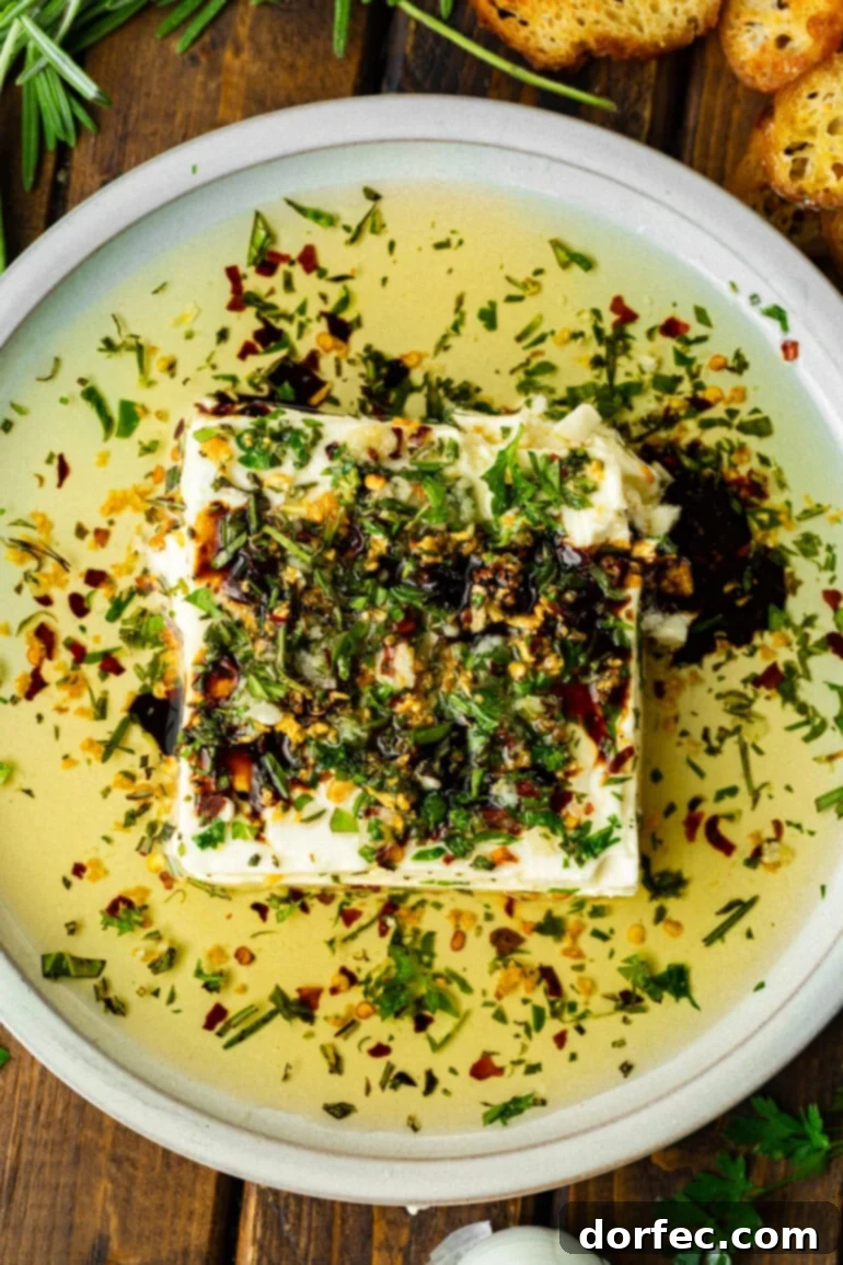 A shallow white circular serving tray presenting a block of Greek feta cheese generously topped with sizzling herb-infused olive oil, ready to be enjoyed with crostini.