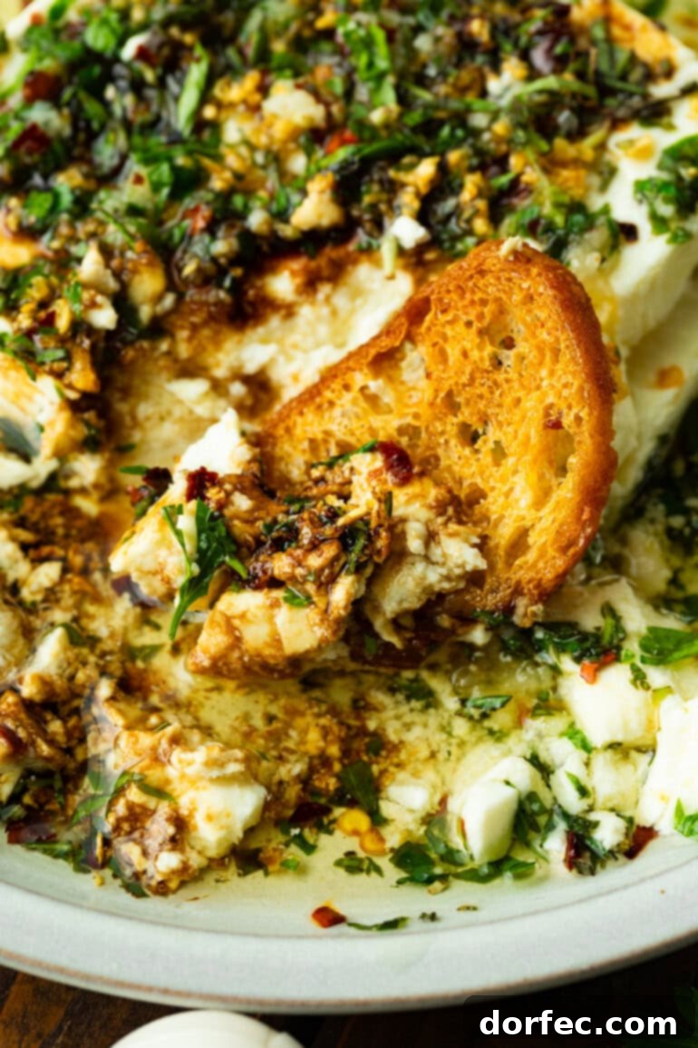 A close-up photo of a crunchy crostini dipped into a bowl of creamy Greek feta, topped generously with hot, aromatic herb-infused olive oil, ready to be savored.