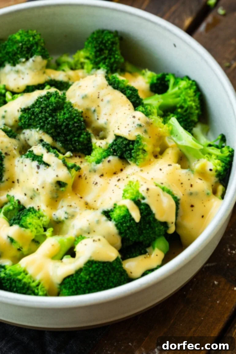 A generous serving of freshly steamed broccoli topped with a rich, creamy cheese sauce and freshly cracked black pepper in a large white bowl.