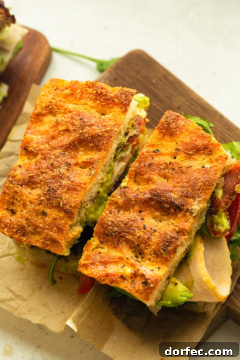 A beautifully presented Turkey Bacon Avocado Sandwich on focaccia bread, meticulously cut in half and resting on a piece of rustic brown parchment paper, ready to be devoured.