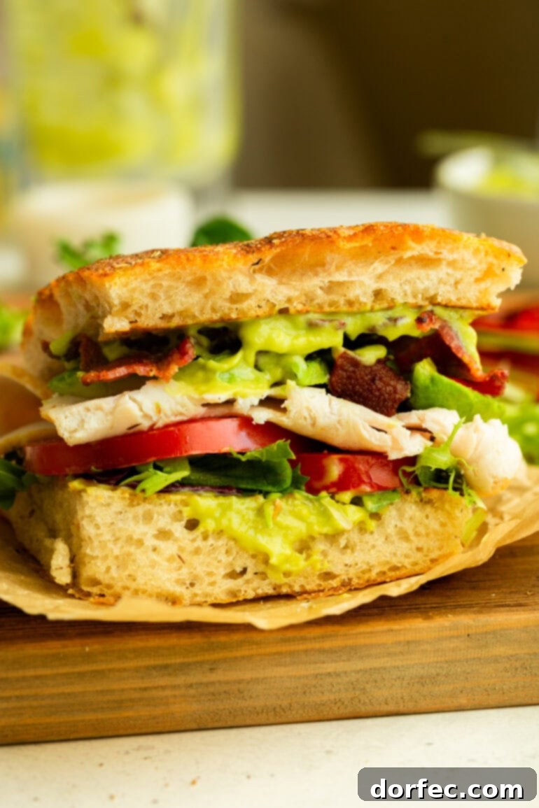A delectable half of a Turkey Bacon Avocado Sandwich, artfully prepared on focaccia bread, showcasing generous layers of honey avocado sauce, fresh tomatoes, mixed greens, thick-cut turkey, and perfectly crispy bacon.