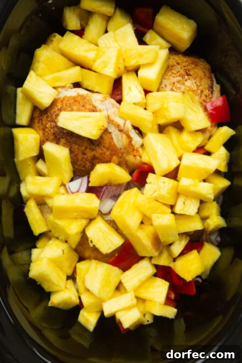 A black crockpot filled with pan-seared chicken thighs, glistening pineapple chunks, vibrant chopped red bell peppers, and thinly sliced purple onion, ready for slow cooking.