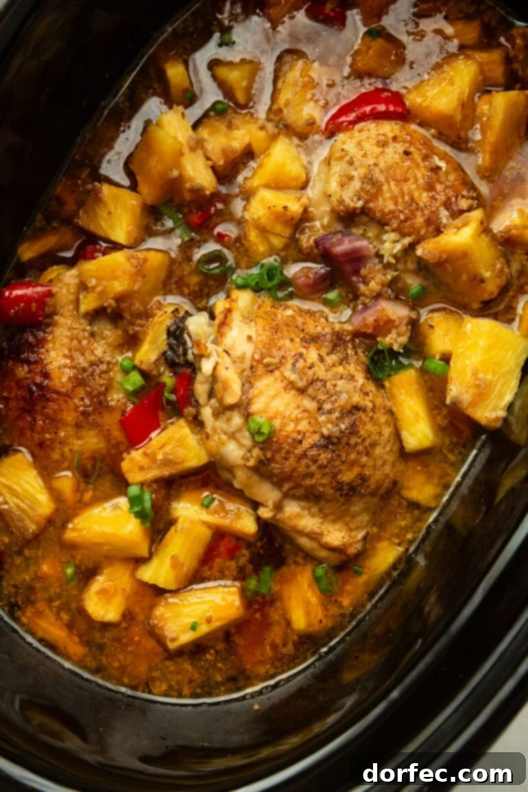 A black crockpot brimming with perfectly cooked chicken thighs, plump pineapple chunks, tender red bell peppers, and softened purple onions, all topped with a vibrant sprinkle of chopped green onions, ready to be served.
