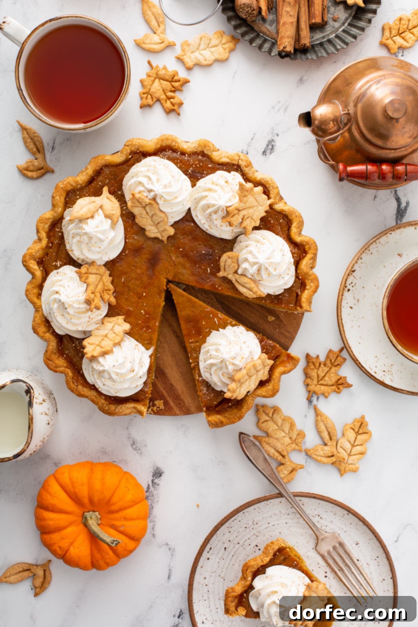 A stunning, golden classic pumpkin pie, beautifully topped with piped swirls of whipped cream and delicate, stamped pie crust leaves, captured from a top-down perspective. A single slice has been carefully removed, showcasing its creamy texture.