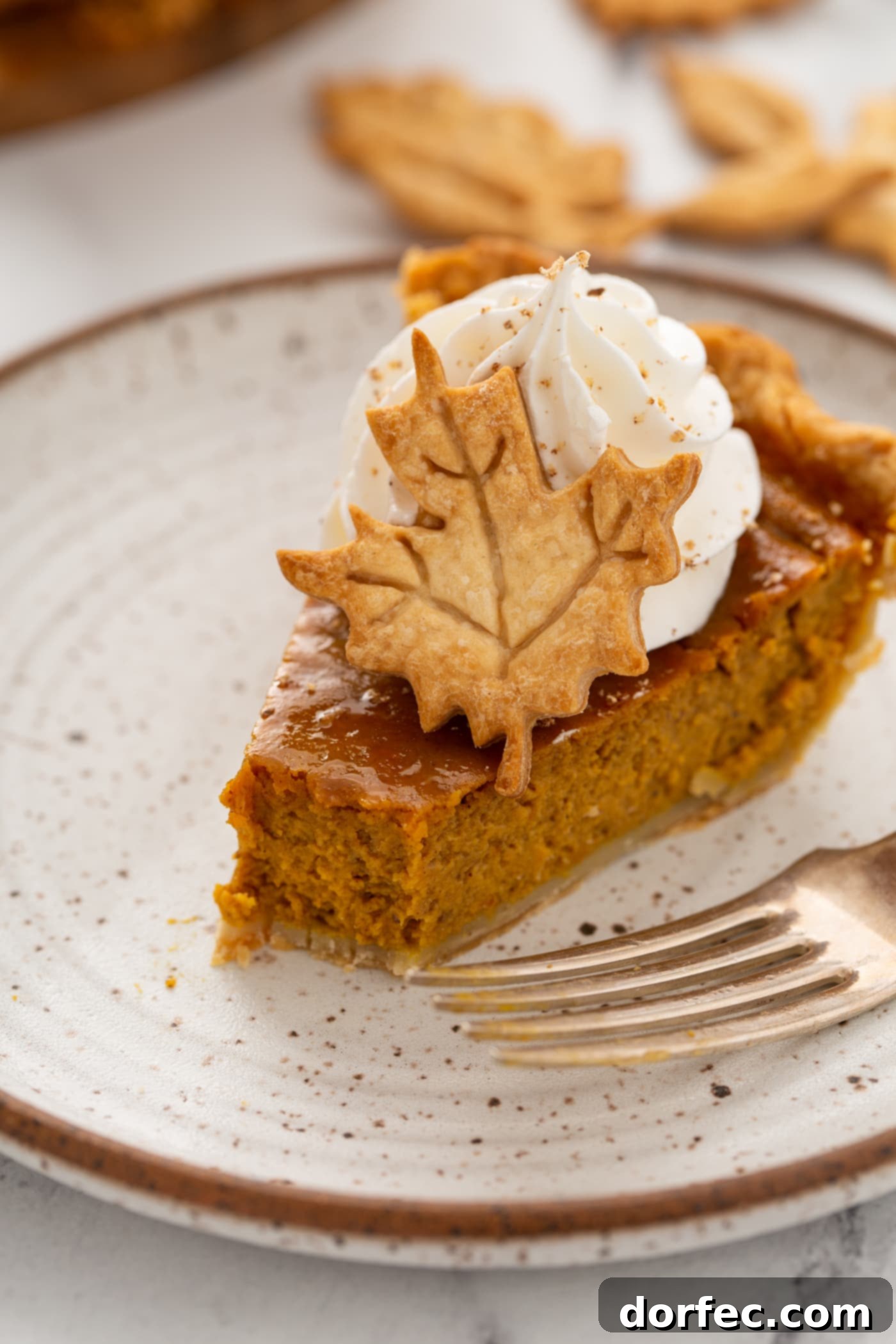 A close-up photo of a perfect slice of homemade pumpkin pie, with a small bite removed, showing its creamy texture. It's topped with piped whipped cream and a delicate pie crust leaf.