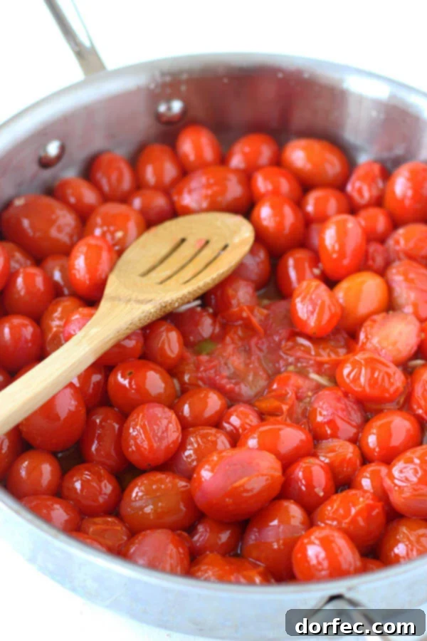 Vibrant grape tomatoes cooking in a pan with a wooden slotted spoon, creating a rich sauce base.