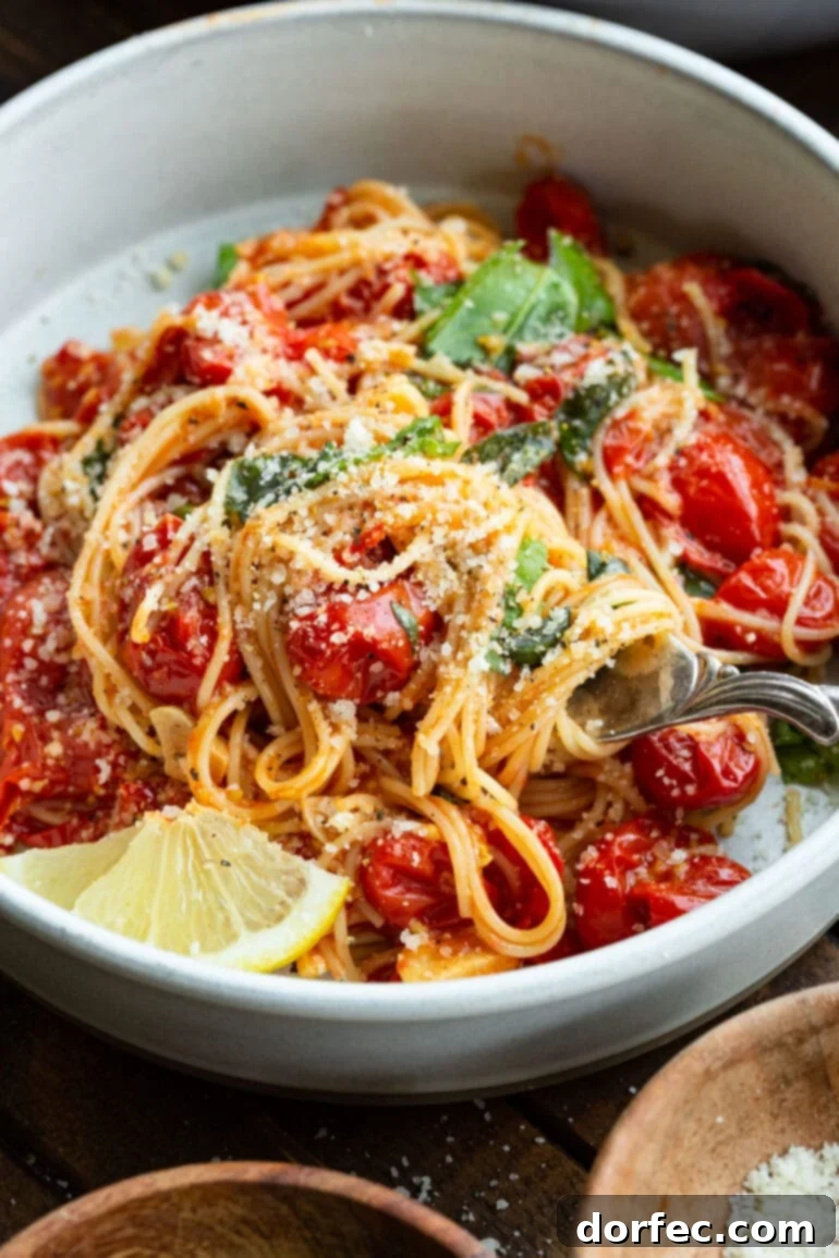 A beautifully presented bowl of angel hair pasta, artfully tossed with cooked grape tomatoes, fresh basil, granulated Parmesan cheese, and garnished with a bright slice of lemon.