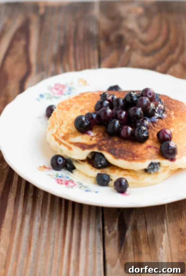 White chocolate blueberry pancakes on ohsweetbasil.com A plate of freshly cooked White Chocolate Blueberry Pancakes, beautifully arranged with a scattering of blueberries and melted white chocolate