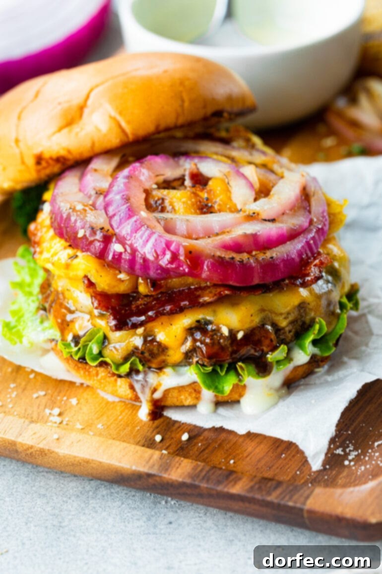 A top-down view of a grilled cheeseburger with the top bun removed, revealing layers of vibrant toppings including thinly sliced red onion, a glistening grilled pineapple ring, and crispy bacon strips, all resting on a perfectly cooked, cheese-covered patty.