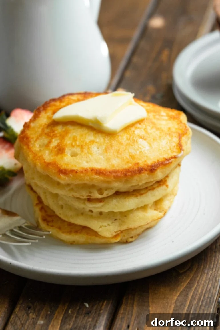 A stack of four golden, fluffy buttermilk pancakes on a white plate, topped with a rich pat of butter, emphasizing their inviting texture and deliciousness for a perfect breakfast.
