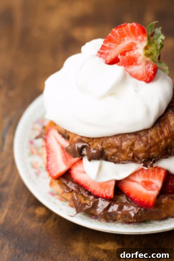 A close-up of a golden grilled croissant generously filled with chocolate hazelnut spread, fresh sliced strawberries, and a dollop of whipped cream, on a plate.