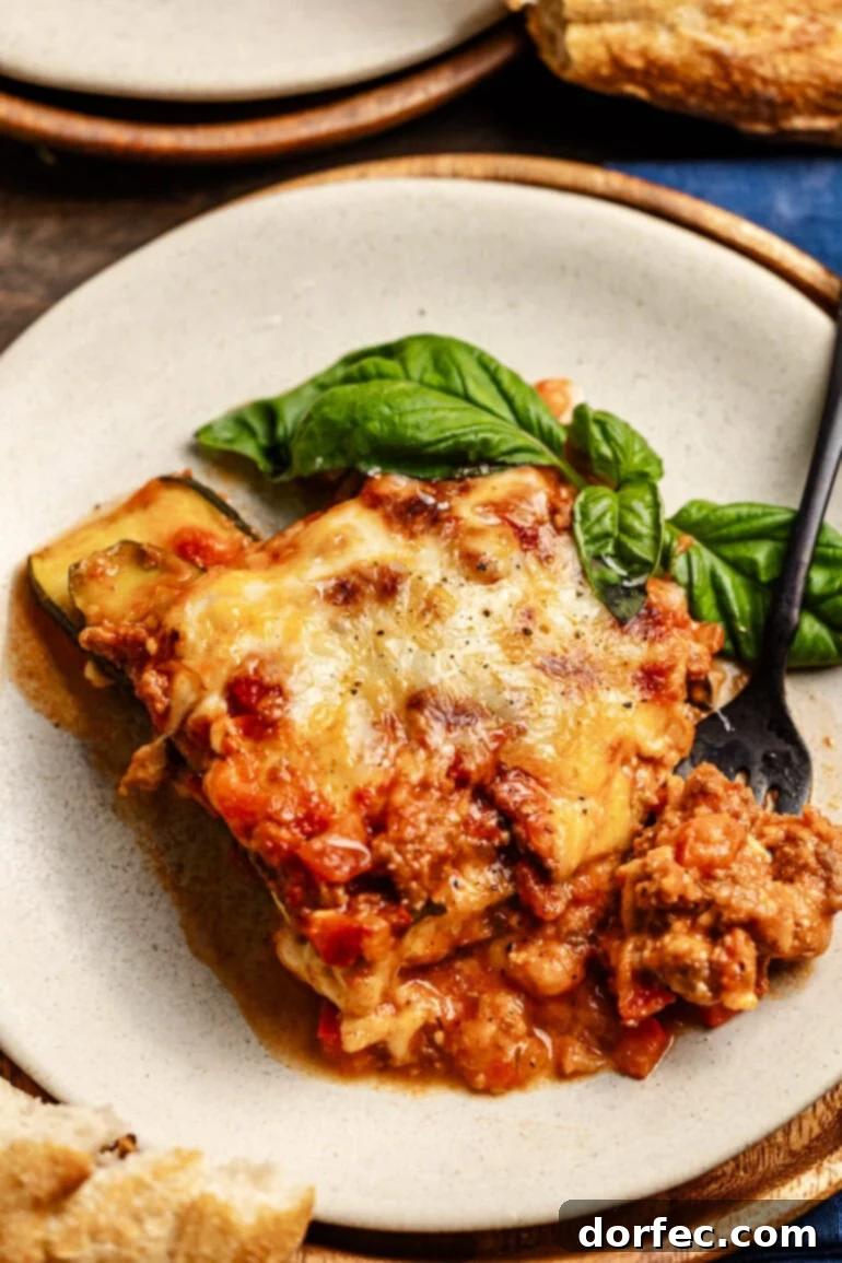A plate with a hearty slice of layered zucchini noodles, cheese, and meat sauce with a basil garnish. A fork is digging into the slice of lasagna, highlighting its rich texture.